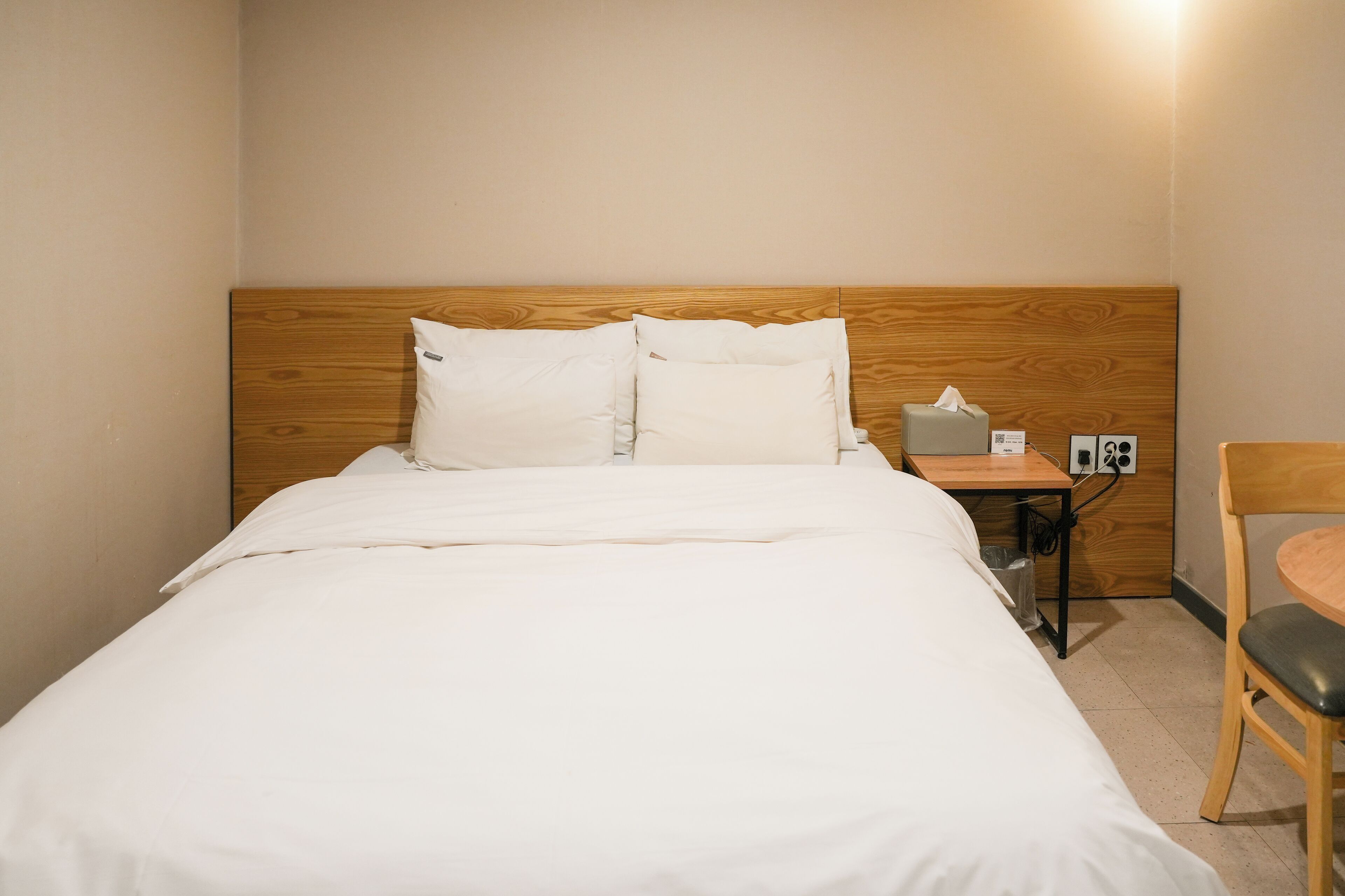 A neatly made bed with white linens, a wooden headboard, a bedside table with a tissue box, and a chair with a round table.