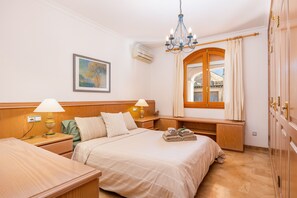 Ein Hotelzimmer mit einem Bett, Nachttischen, einer Lampe, einem Gemälde, einer Klimaanlage, einem Fenster mit Vorhängen und einer Chandelieber.