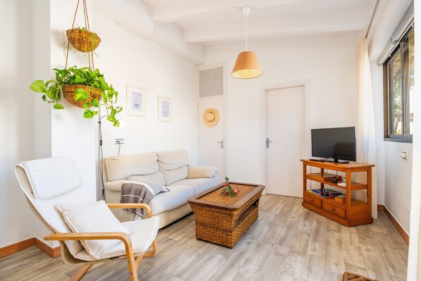 Un salón con un sofá, una silla, un televisor sobre un mueble de madera y dos cestas de mimbre con plantas.