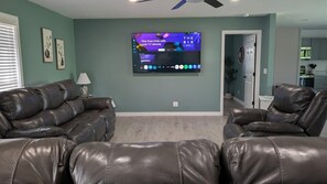 Uma sala de estar com televisão na parede, dois sofás de couro, um ventilador de teto e uma porta que dá para uma cozinha.