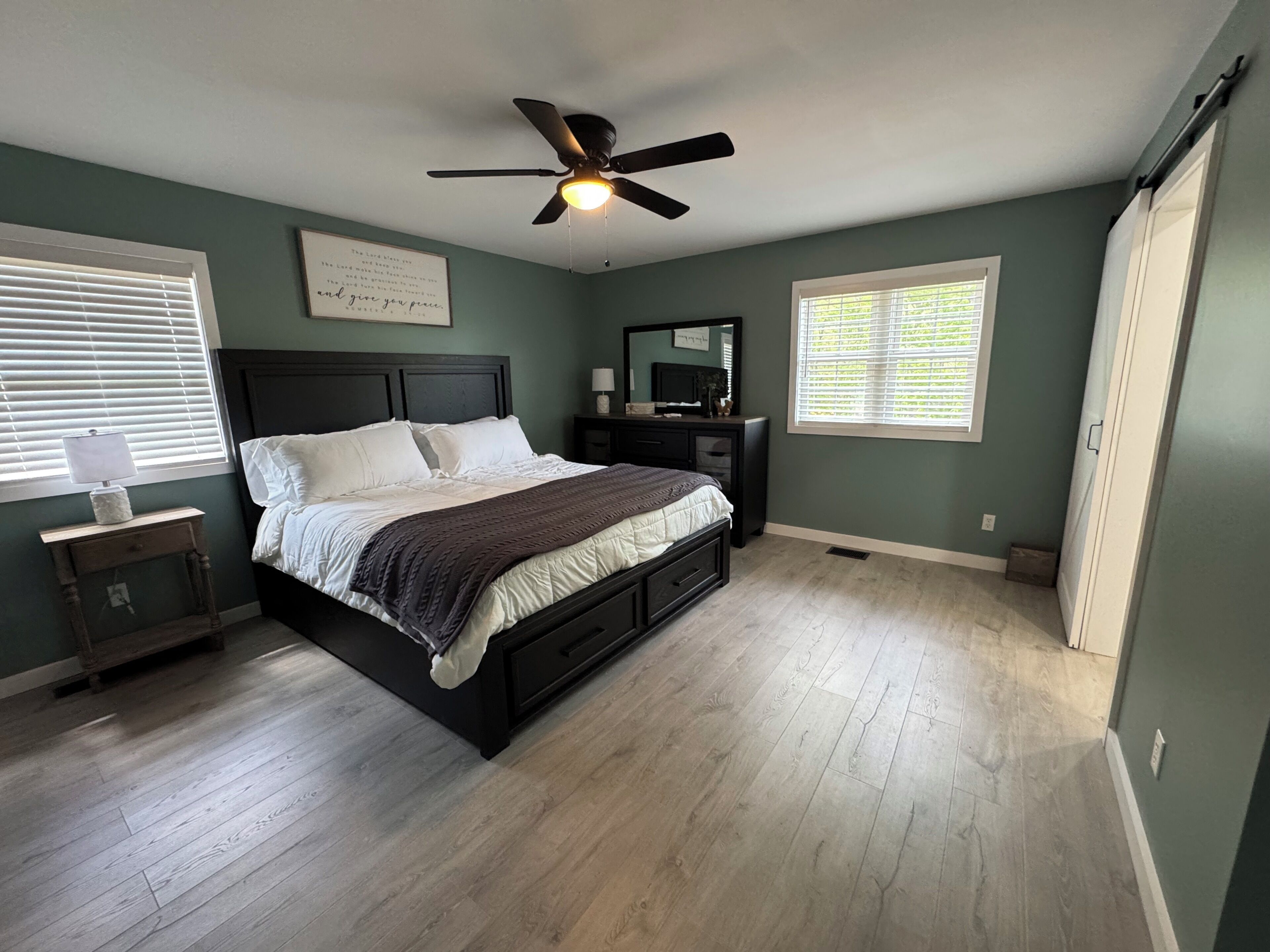 Une chambre à coucher comprenant un lit, un ventilateur de plafond, une commode, une table de chevet et une fenêtre avec des stores.