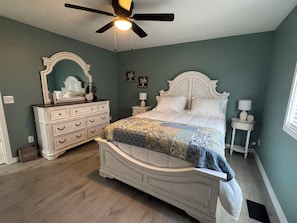 Quarto com cama, penteadeira, mesas de cabeceira, ventoinha no teto e janela.