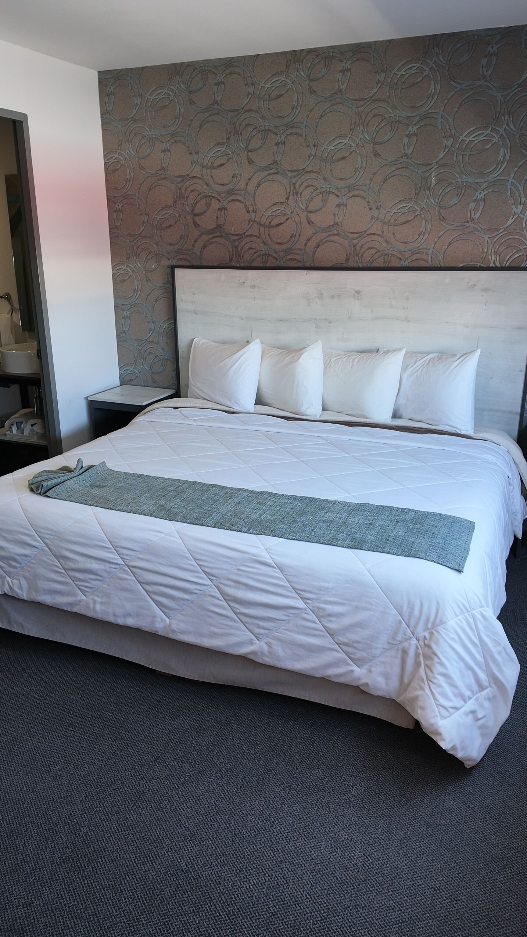 A hotel room with a large bed, a headboard with a circular pattern, a grey throw blanket, and a dark carpet.