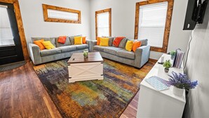 A living room with two grey sofas, colorful cushions, a white coffee table, a multicolored rug, and wooden-framed windows.
