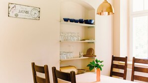 Een eetruimte met een witte tafel, houten stoelen, een hangende hanglamp, planken met blauwe kommen en drinkglazen, en een bordje aan de muur.