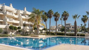A swimming pool with a clear blue water surface, surrounded by a tiled deck. In the background, there are multi-story buildings with balconies and palm trees. - Palm Breeze Retreat in Albufeira w/ Pool & Balcony (Albufeira)