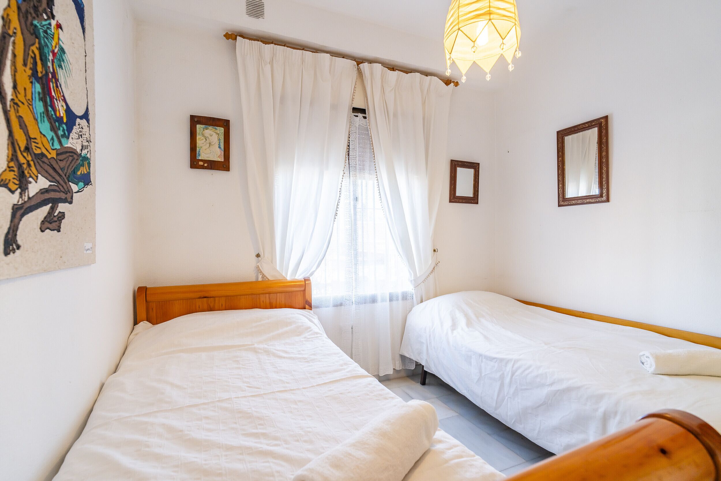 Una stanza con due letti, biancheria da letto bianca, una testiera di legno, una finestra con tende trasparenti, un quadro appeso al muro e una lampada a sospensione.
