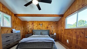 Camera da letto con pareti in legno, un letto con copriletto grigio, due comodini con lampade, una ventola a soffitto e una finestra con vista sugli alberi.