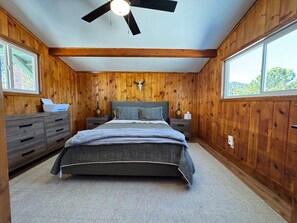 A bedroom with wooden walls, a bed with a gray quilt, two bedside tables with lamps, a ceiling fan, and a window with a view of trees.
