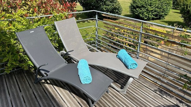 Two sun loungers with towels on a wooden deck with a metal railing, surrounded by greenery.
