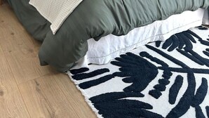 A neatly made bed with a green and white duvet, a rolled towel, and a black and white patterned rug on a wooden floor.