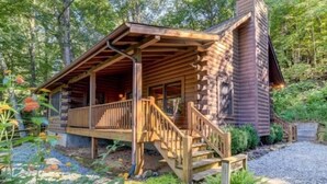 Une cabane en rondins dotée d’un porche, d’un escalier en bois et d’une allée gravillonnée, le tout entouré de verdure.