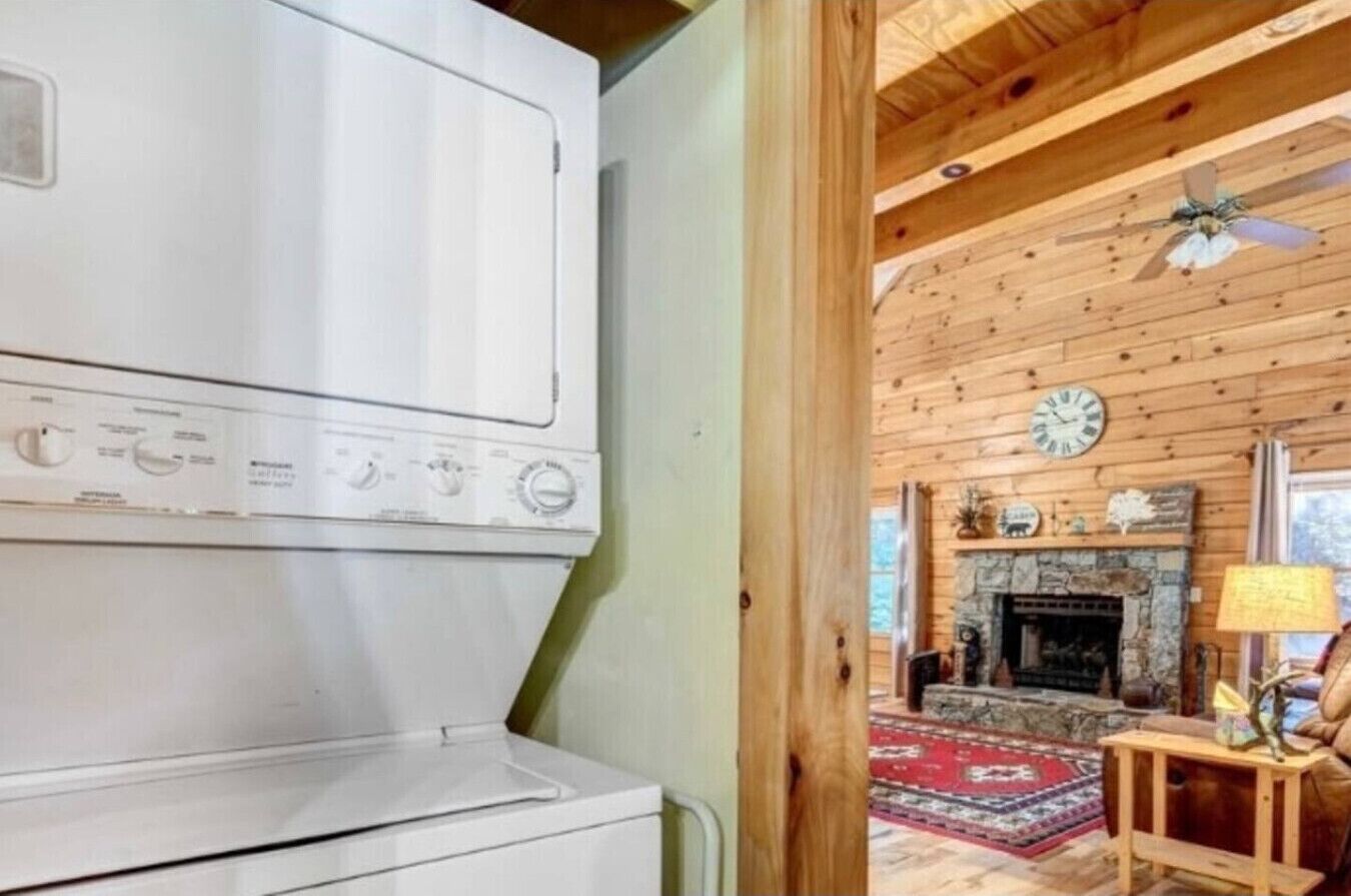 A laundry room with a large white washer and dryer, a wooden ceiling, and a rustic interior with a fireplace and a clock on the wall.