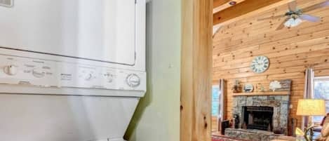 A laundry room with a large white washer and dryer, a wooden ceiling, and a rustic interior with a fireplace and a clock on the wall.