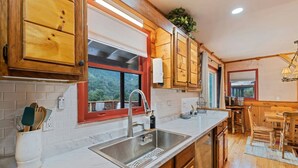 Una cocina con armarios de madera, un lavamanos de acero inoxidable y un ventanal con marco rojo.