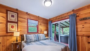 Un dormitorio con paneles de madera, una cama, dos mesitas de noche con lámparas, una ventana con cortinas y vistas al verdor exterior.