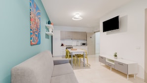 Ein modernes Wohnzimmer mit einer Couch, einem Esstisch mit Stühlen, einer Küche im Hintergrund und einem an der Wand befestigten Fernseher.