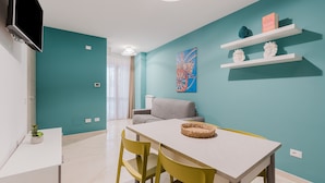 Ein modernes Wohnzimmer mit türkisfarbenen Wänden, einem weißen Tisch, gelben Stühlen, einem grauen Sofa und einem Wand-Fernseher.