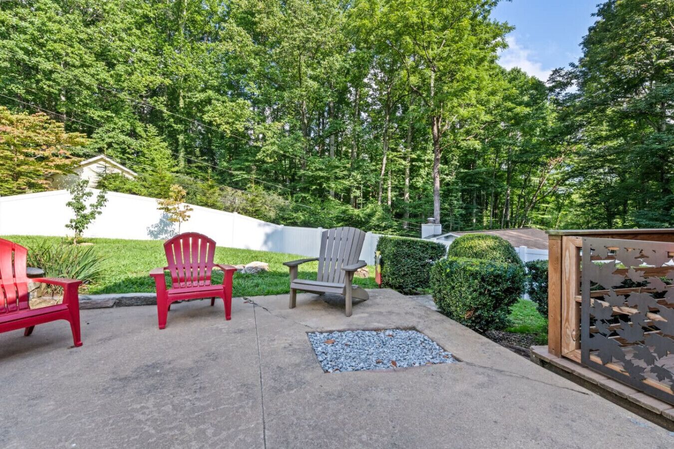 An outdoor patio with three Adirondack chairs, a wooden fence, and a hedge.