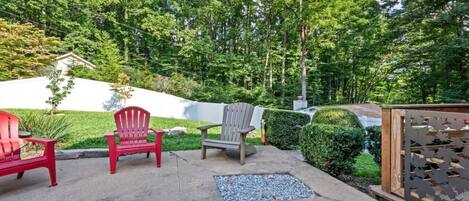 An outdoor patio with three Adirondack chairs, a wooden fence, and a hedge.