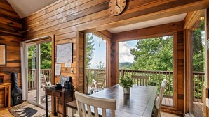 Interior de una cabaña de madera con una mesa de comedor, sillas, un reloj y vistas al exterior.