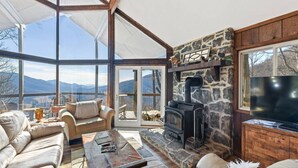 Ein geräumiges Wohnzimmer mit Kamin aus Stein, bequemen Sitzgelegenheiten und einem großen Fenster mit Blick auf die Berge.