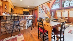 A spacious kitchen with a stone wall, wooden cabinets, and a dining area with a wooden table and chairs. - Mountain Laurel-Secluded Beauty + Majestic Views! (Maggie Valley)