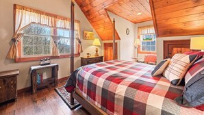 A cozy bedroom with a wooden ceiling, a large bed with a checkered blanket, a nightstand, a window with curtains, and a small table with books. - Mountain Laurel-Secluded Beauty + Majestic Views! (Maggie Valley)