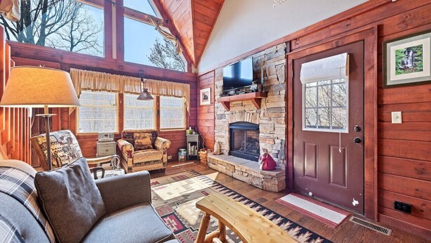 A cozy cabin interior with a stone fireplace, wooden walls, a sofa, and a large window. - Mountain Laurel-Secluded Beauty + Majestic Views! (Maggie Valley)