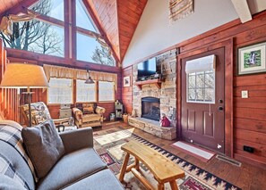 A cozy cabin interior with a stone fireplace, wooden walls, a sofa, and a large window. - Mountain Laurel-Secluded Beauty + Majestic Views! (Maggie Valley)