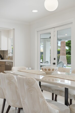 A modern kitchen with white cabinetry, a dining table with a white and beige patterned table runner, and a view of the outdoors through a glass door. - "La Veranda" a Movie Colony Palm Springs Resort Pool and View Home, #069334 (Palm Springs)