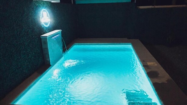 Una piscina rectangular con una fuente de agua azul claro, rodeada por un muro oscuro y una estructura pequeña e iluminada.