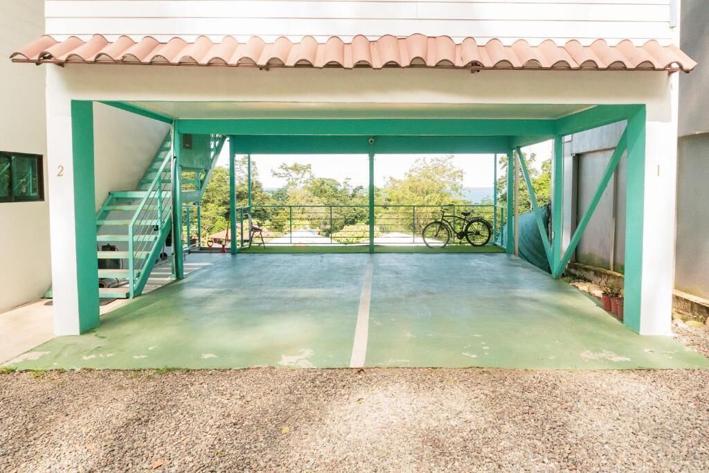Ein überdachter Außenbereich mit einer Treppe, die zu einer zweiten Etage führt, ein abgestelltes Fahrrad und im Hintergrund Grünflächen sowie ein Gebäude.