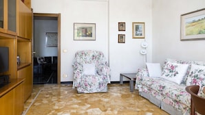 Uma sala de estar com um sofá, uma poltrona estampada, uma televisão e um armário de madeira.