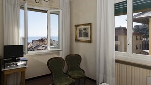 Quarto de hotel com uma cama, uma secretária com televisão, duas cadeiras, uma janela com vista para edifícios e o mar, e um quadro na parede.
