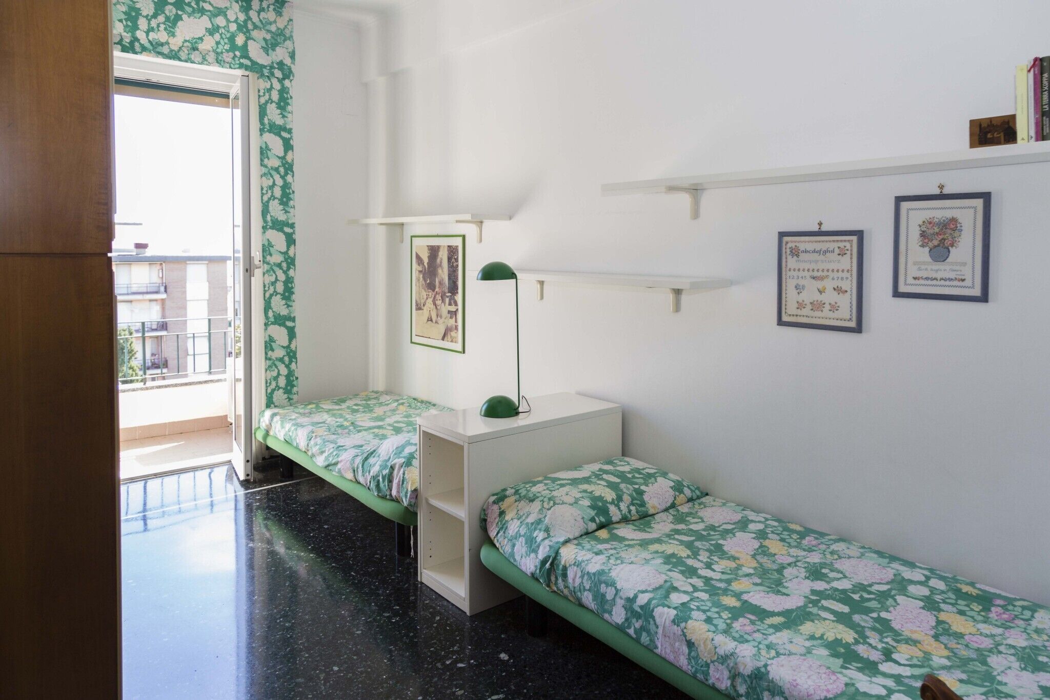 Una habitación con dos camas individuales, una mesita de noche pequeña y blanca con una lámpara verde, un cuadro en la pared y un balcón con vistas.