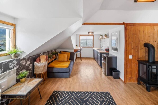 Een kamer in loftstijl met een bank, eettafel, keukenhoek en een houtkachel.