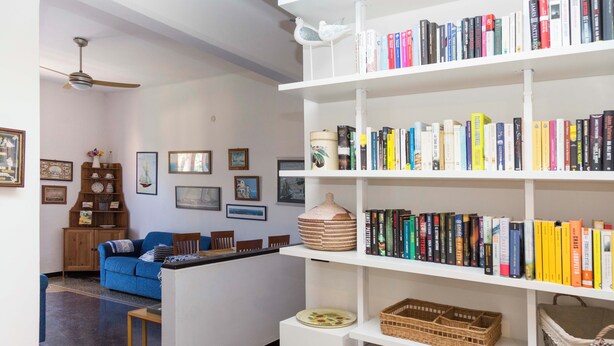 Una sala de estar con un sofá azul, una estantería blanca llena de libros y un ventilador de techo.