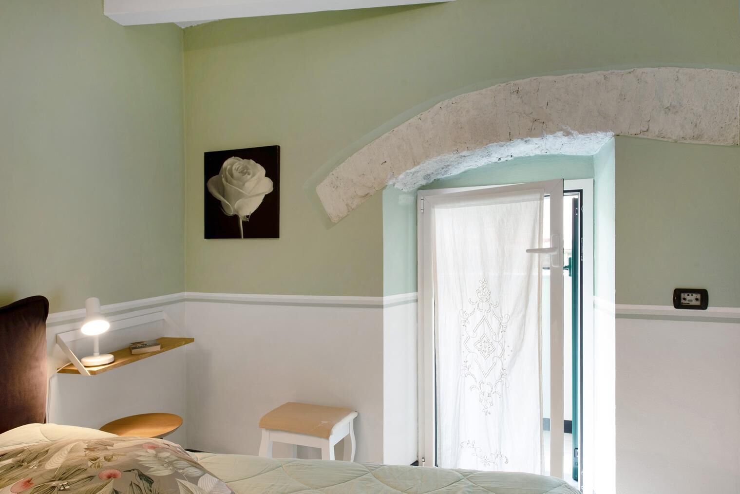 Ein Schlafzimmer mit Bett, Nachttisch, Schreibtischlampe, einem kleinen Hocker, einem gerahmten Bild und einer Tür mit Vorhang.