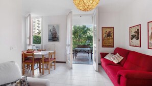 Ein helles Wohnzimmer mit einem roten Sofa, einem Essbereich mit Holzsesseln und einem großen Fenster mit Blick auf Grünflächen.