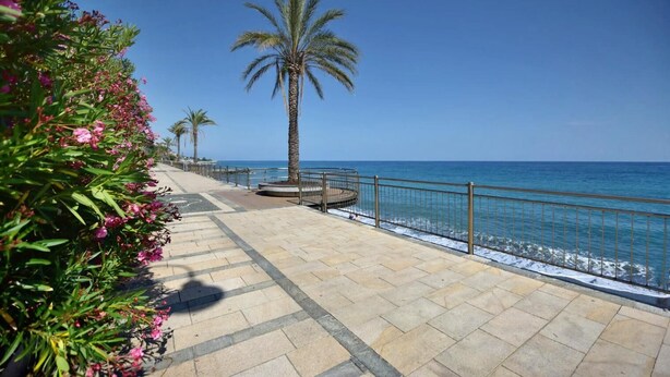 A paved walkway with palm trees and flowers, leading to a beach with a railing. - Casa Luxury Golf - One Bedroom Apartment, Sleeps 3 (Arenzano)