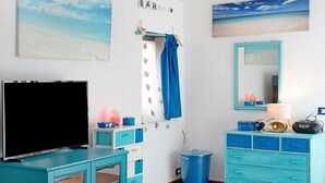 Una stanza con una TV, un cestino blu, un mobiletto con cassetti blu, uno specchio e un quadro raffigurante una spiaggia.