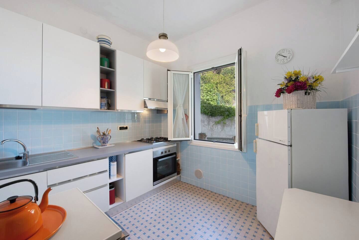 Une cuisine compacte avec des meubles blancs, une cuisinière à gaz, un évier et une fenêtre donnant sur un espace verdoyant.