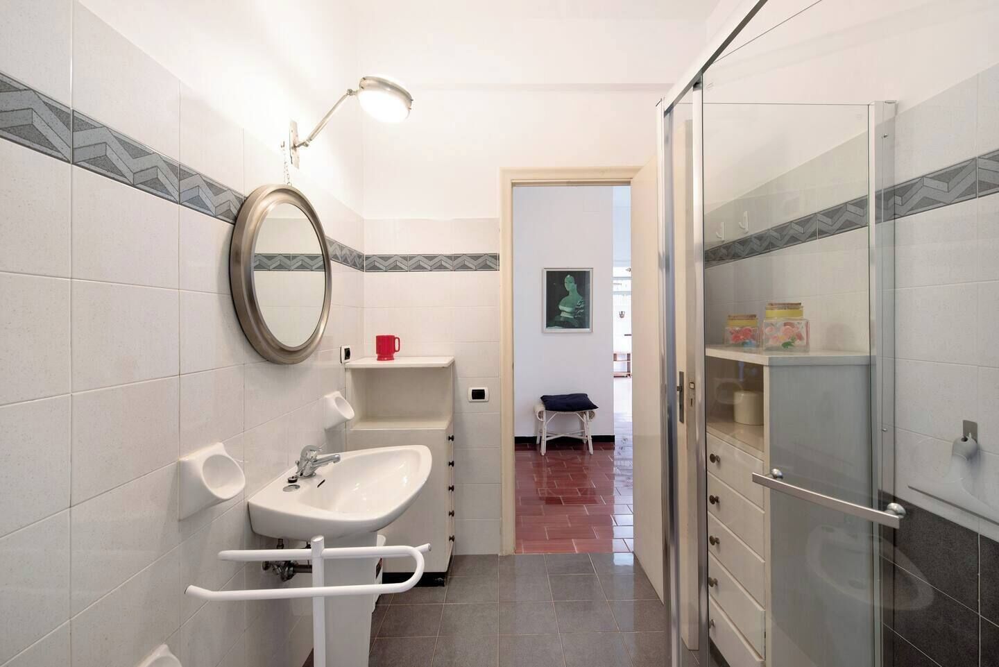 Un baño moderno con lavamanos, espejo y ducha. Hay una silla y una estantería con objetos.