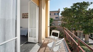 A balcony with a white chair, a door leading to a room with a bed, and a view of buildings and trees. - Casa Olivette - Two Bedroom Apartment, Sleeps 5 (Arenzano)