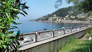 Ein Küstenweg mit einer Betonbarriere, der auf einen Strand mit Menschen und Sonnenschirmen blickt.