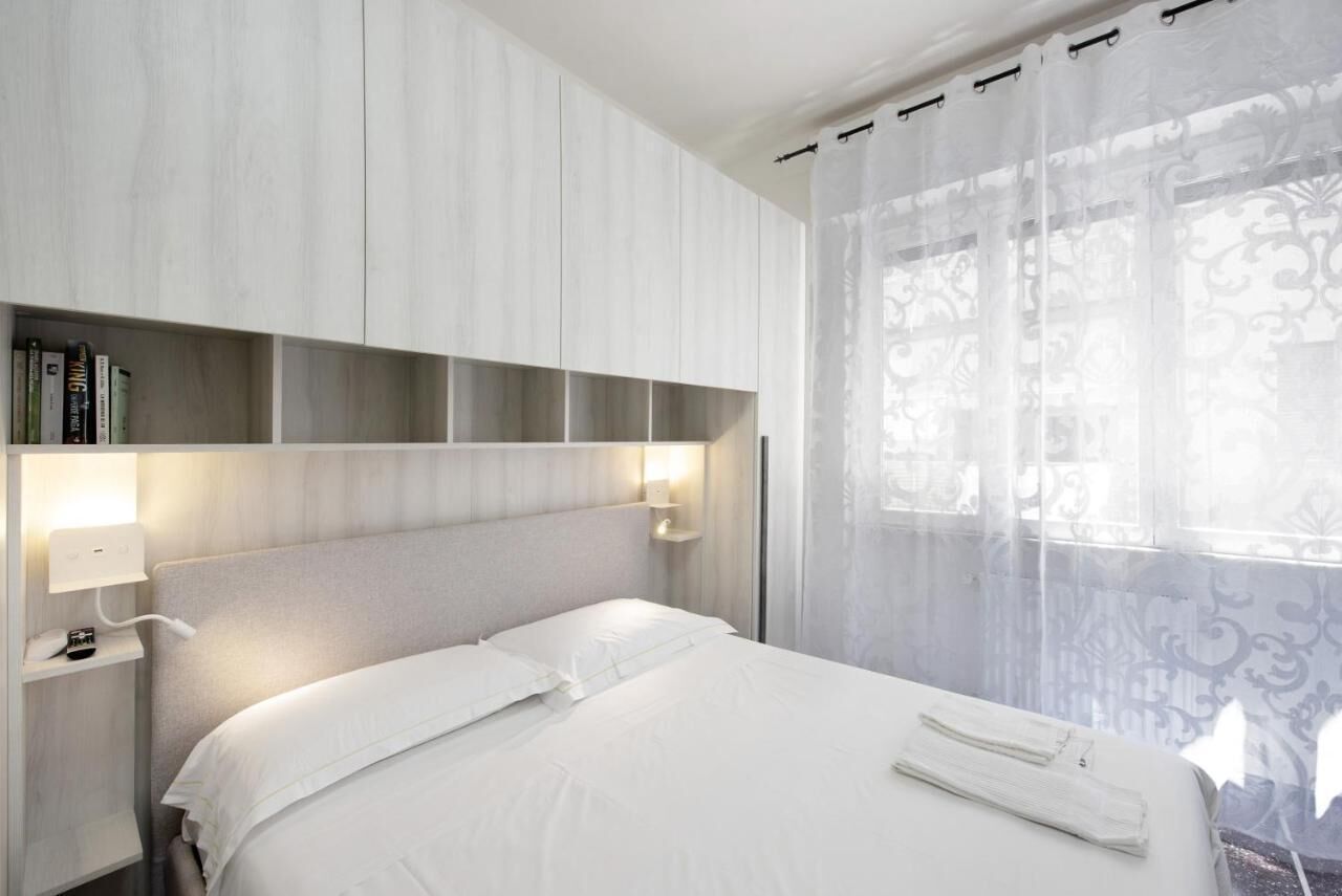 Una habitación de hotel con cama, lámparas de noche, estantería y ventana con cortinas translúcidas.