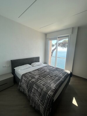 A modern bedroom with a large bed, a view of the sea through a sliding glass door, and a minimalist design. - L'Ile d'Azur - Three Bedroom Villa, Sleeps 6 (Arenzano)