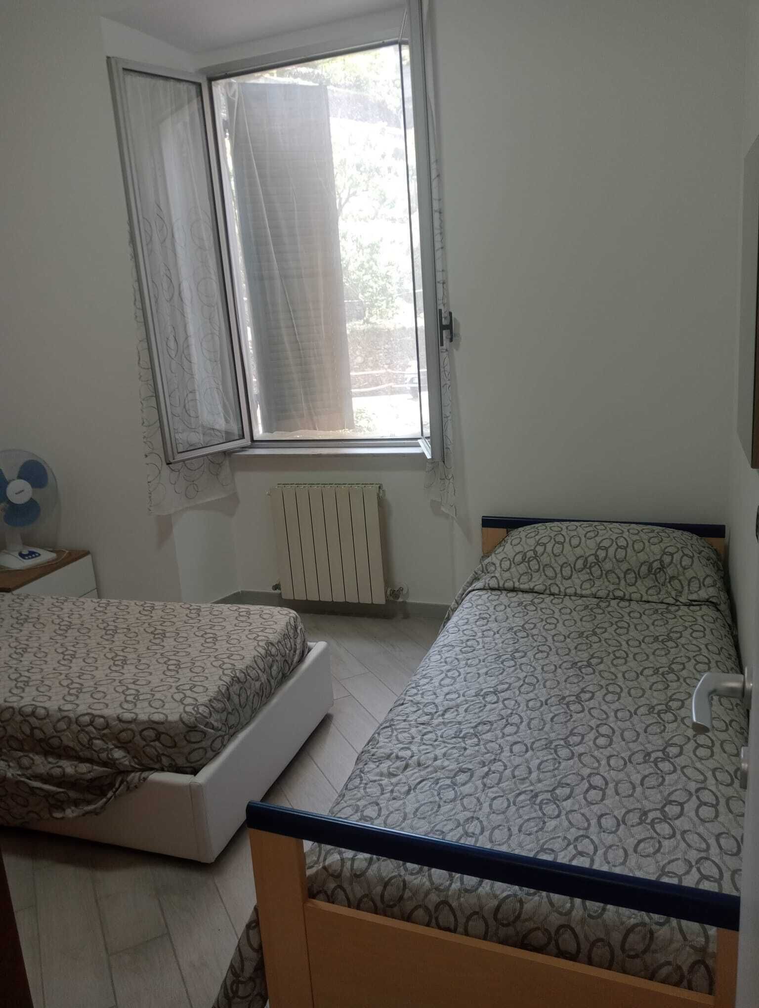 A bedroom with two beds, a window with curtains, a radiator, and a bedside table with a fan.