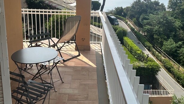Varanda com mesa e cadeiras, guarda-corpo branco e vista para uma estrada e vegetação.